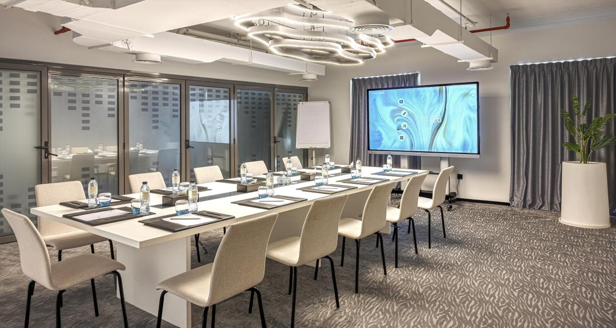 Апарт готель Millennium Atria Business Bay Дубай Екстер'єр фото The photo shows a modern conference room set up for a meeting. There is a long table with several chairs arranged around it, each with a notepad and pen. A large screen displays a graphic or presentation, and there is a whiteboard in the corner. The 