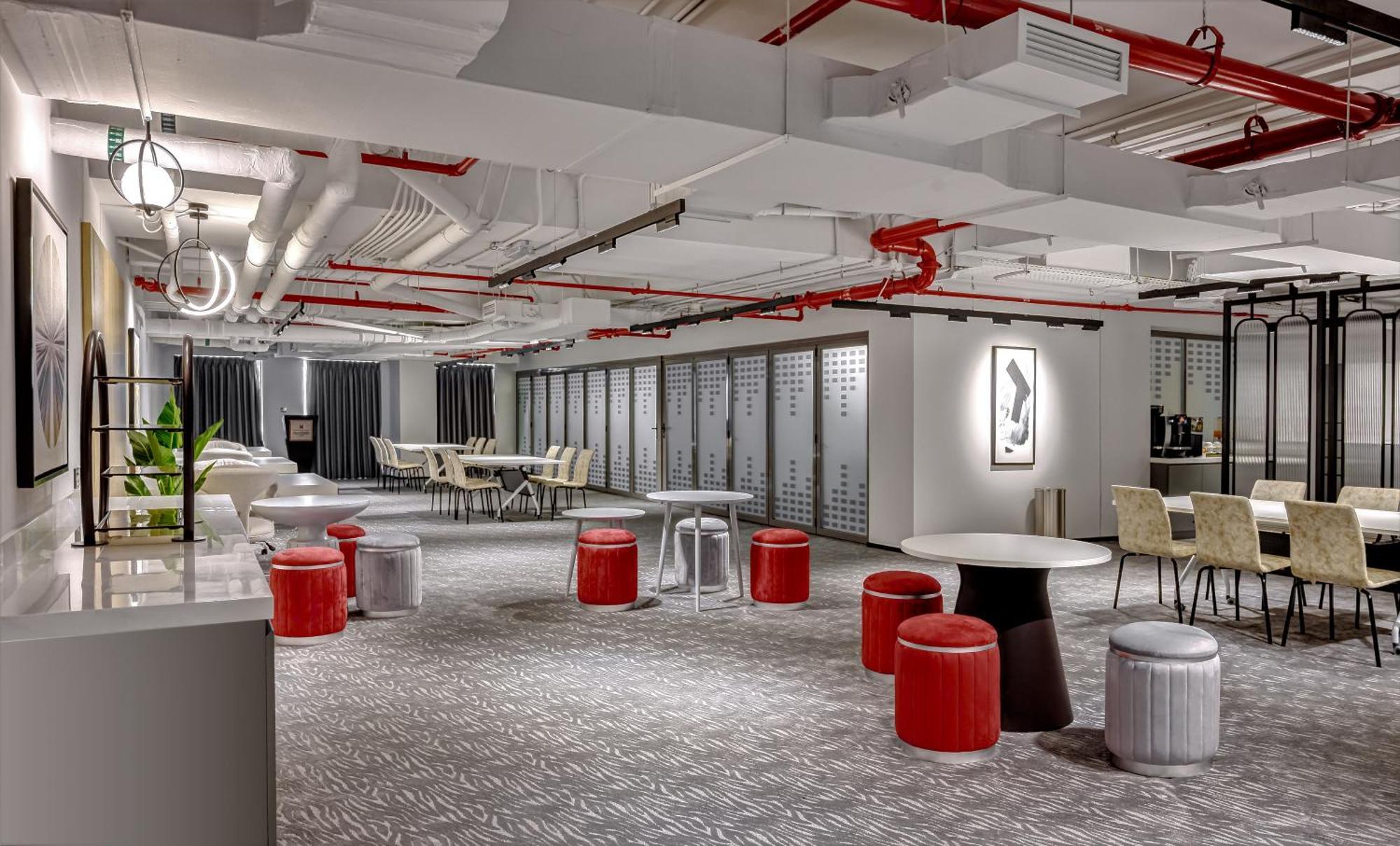 Апарт готель Millennium Atria Business Bay Дубай Екстер'єр фото The photo shows a modern interior space, likely an office or communal area. The flooring is covered with a textured carpet, and the ceiling features exposed pipes painted red. 

There are several seating options, including small round tables with sto