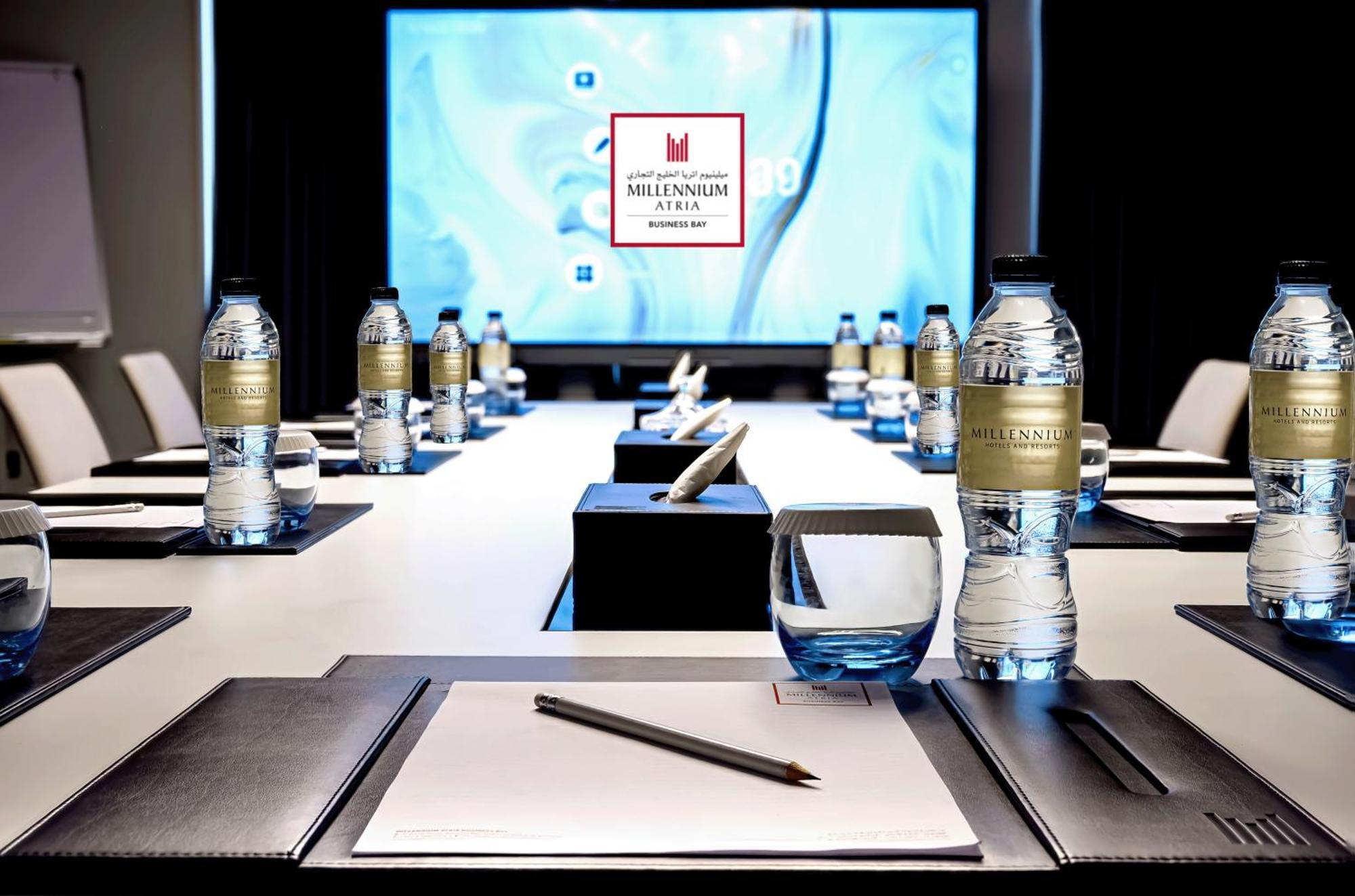 Апарт готель Millennium Atria Business Bay Дубай Екстер'єр фото The photo shows a modern conference room setup. A long table is laid out with neatly arranged stationery, including notebooks and pens, at each seat. Several bottles of water are placed on the table. In the background, a large screen is displayed, li