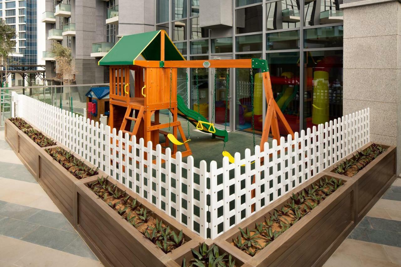 Апарт готель Millennium Atria Business Bay Дубай Екстер'єр фото The image shows a children's playground area, which is enclosed by a white picket fence. The playground features a wooden play structure with a slide, swings, and climbing elements. Surrounding the play area are planters with greenery, and the backdr