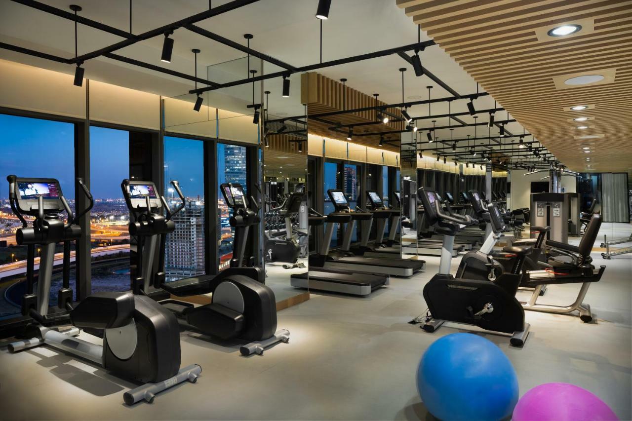 Апарт готель Millennium Atria Business Bay Дубай Екстер'єр фото The photo shows a modern gym with various types of exercise equipment available. There are several treadmills and elliptical machines arranged in rows, with large windows in the background providing a view of the city. The lighting is bright and cont