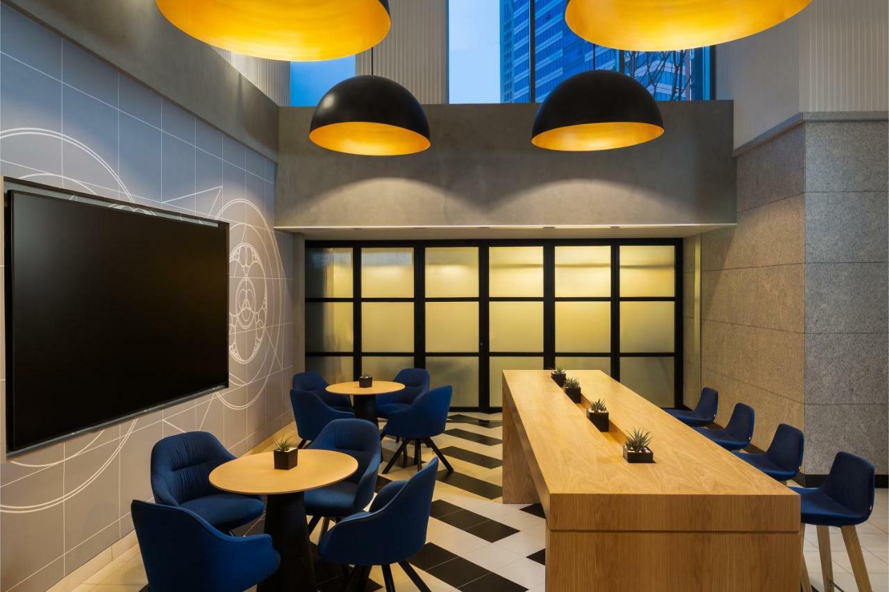 Апарт готель Millennium Atria Business Bay Дубай Екстер'єр фото The photo shows a modern interior space, likely a lounge or meeting area. It features a long wooden table with some small plants on it, accompanied by several blue, circular chairs arranged around it. There is a large flat-screen television mounted o