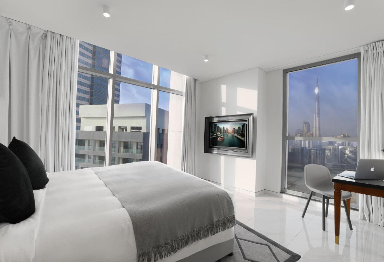 Апарт готель Millennium Atria Business Bay Дубай Екстер'єр фото The photo depicts a modern hotel room that features a large bed with a gray throw blanket. There are large windows allowing natural light to fill the space, showcasing a city skyline view. A flat-screen television is mounted on the wall opposite the 